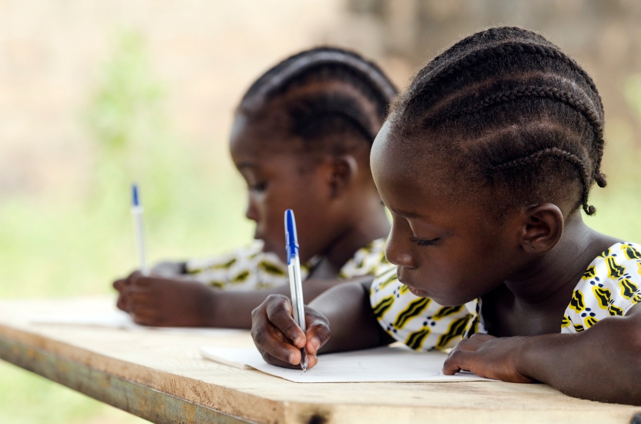 African Children at School Doing Homework. African ethnicity students writing their essay in an African school. They're holding blue pens to write down their homework whilst sitting in their desk.